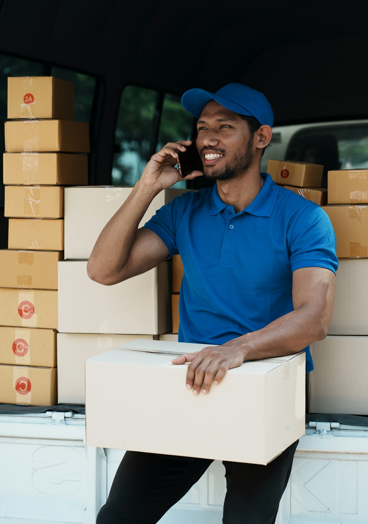 Delivery man with smartphone and delivery box, sitting on the van and contact with customer.
